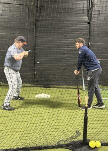 Hunter Weissman leans on a bat while giving instruction to Rob Thayer on his turn and posture mechanics